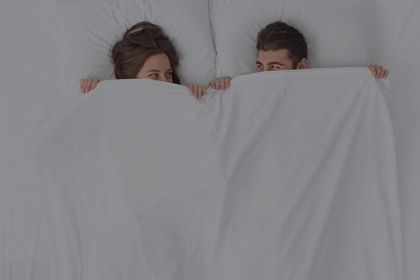 Playful couple peeking out from under a white duvet